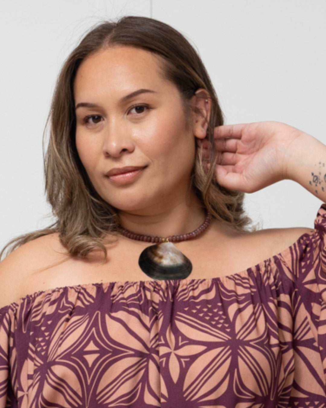 Woman wearing a patterned off-shoulder top with a necklace against a plain background