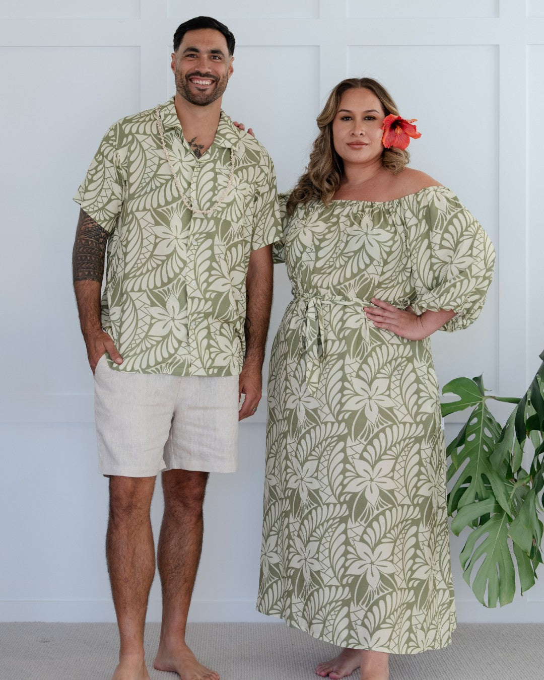 Man and woman wearing matching green leaf-patterned island wear standing against a white background.