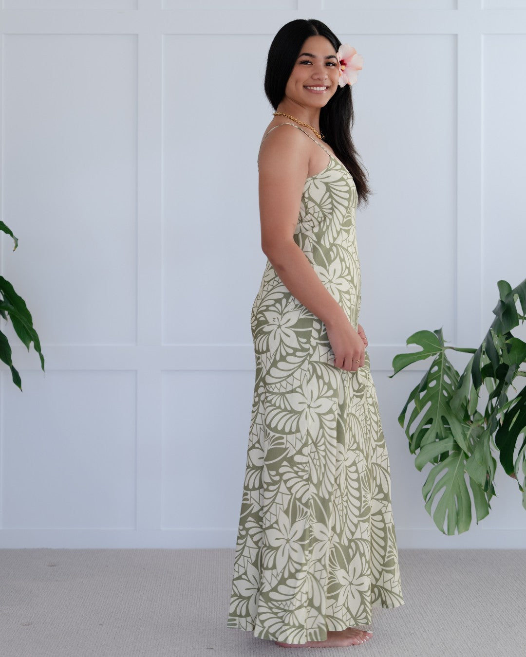Woman wearing a long floral island dress against a white wall with plants.