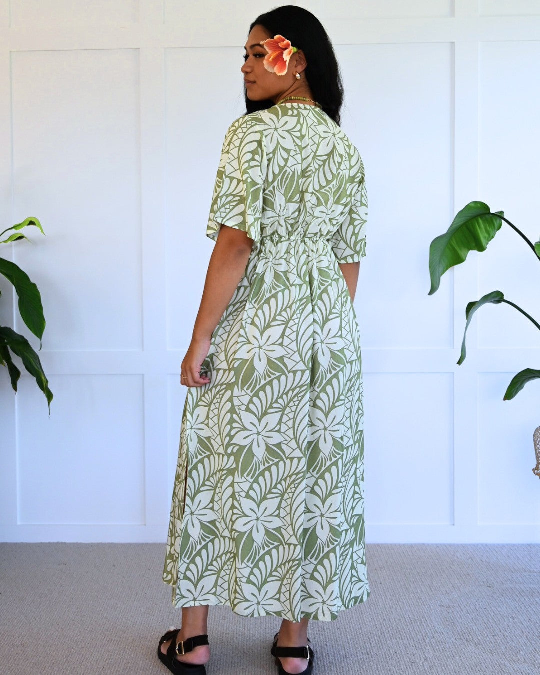 Woman wearing a green and white patterned island dress standing in a room with plants.