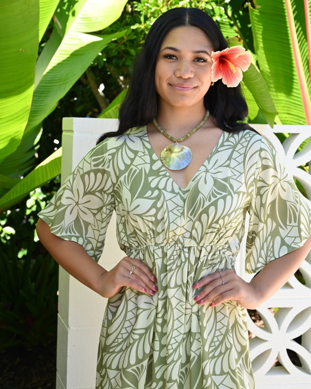 Woman wearing a island floral dress with a flower in her hair, standing among green foliage.