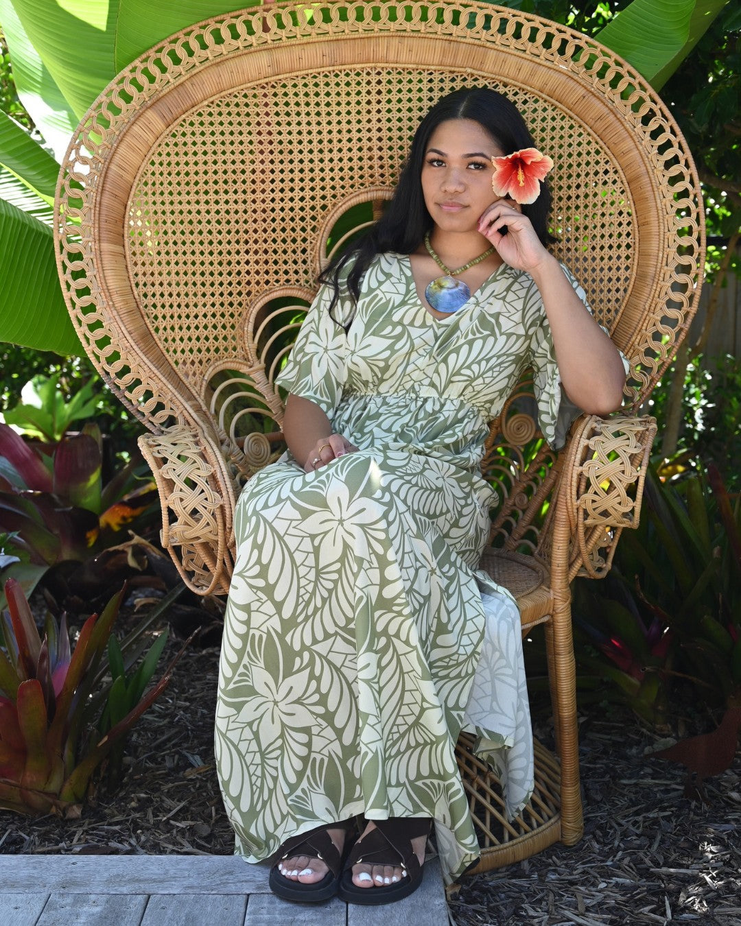 Woman wearing a island floral dress with a flower in her hair, standing among green foliage.