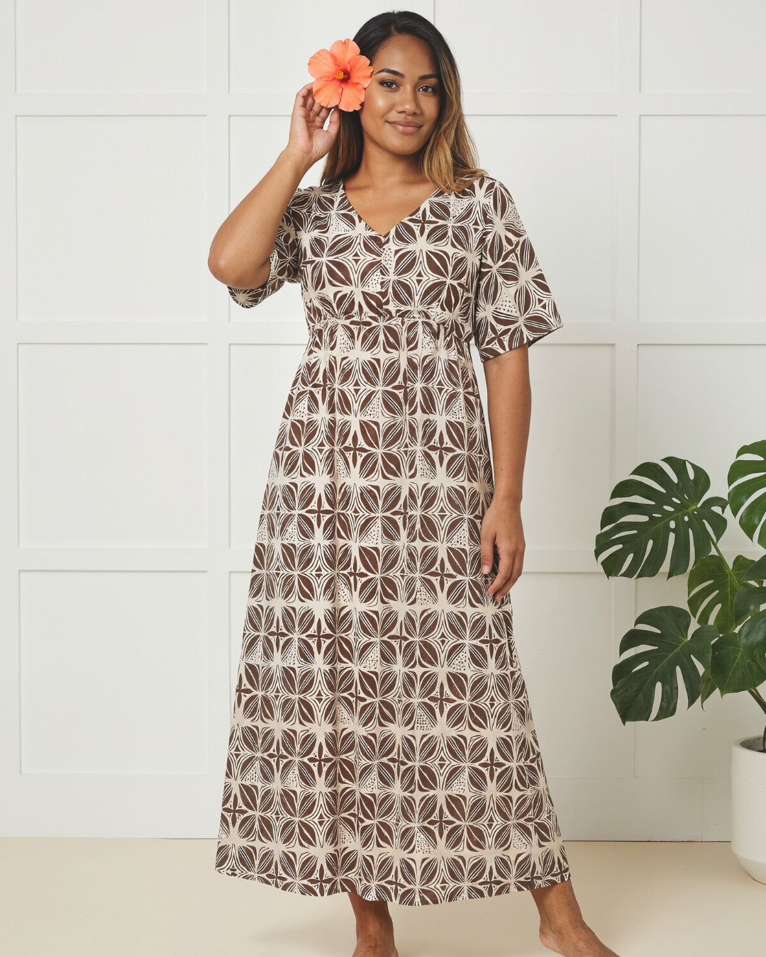 Woman wearing a patterned dress holding an orange flower in a minimal indoor setting.