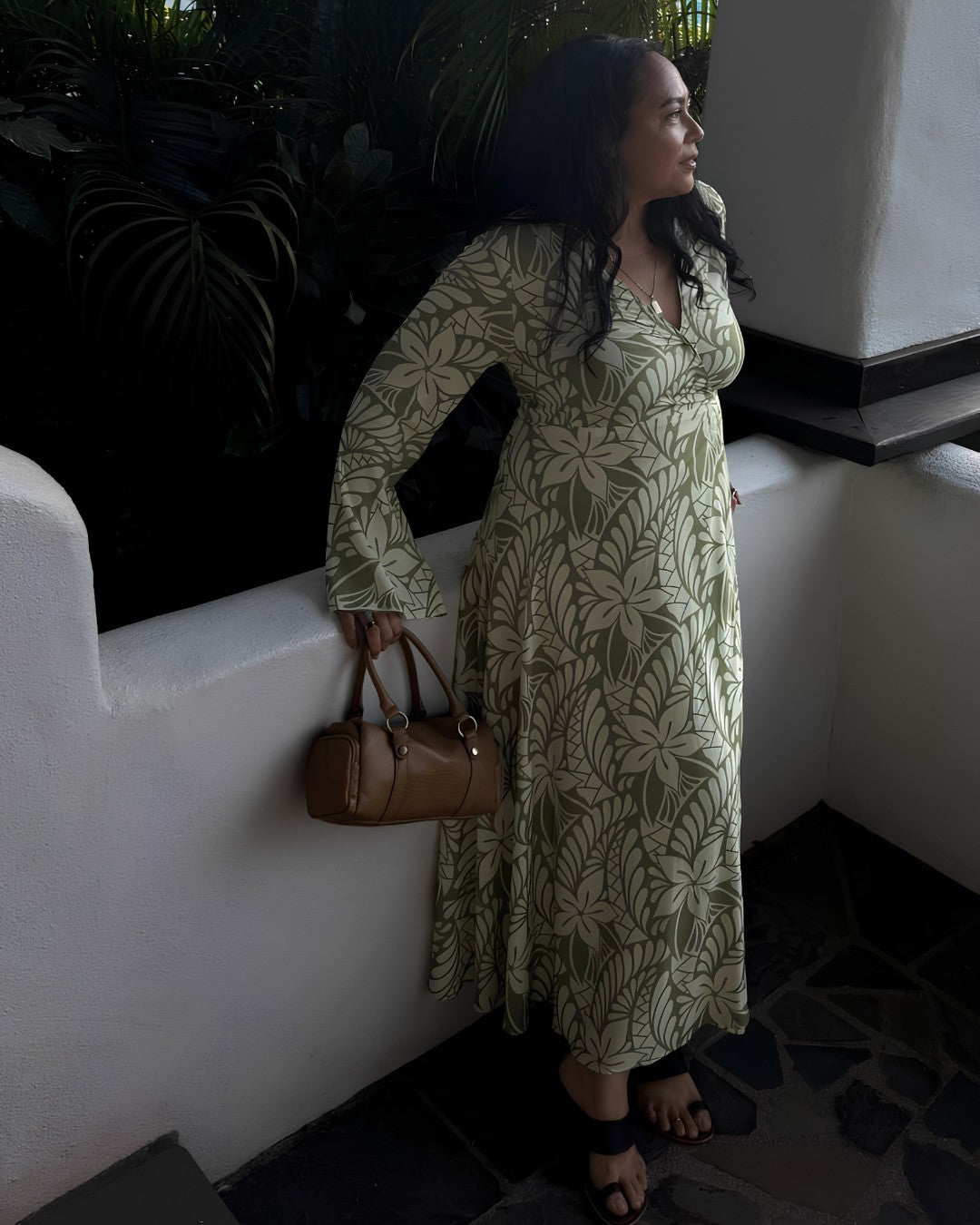 Woman in a green floral dress holding a brown handbag against a white wall.