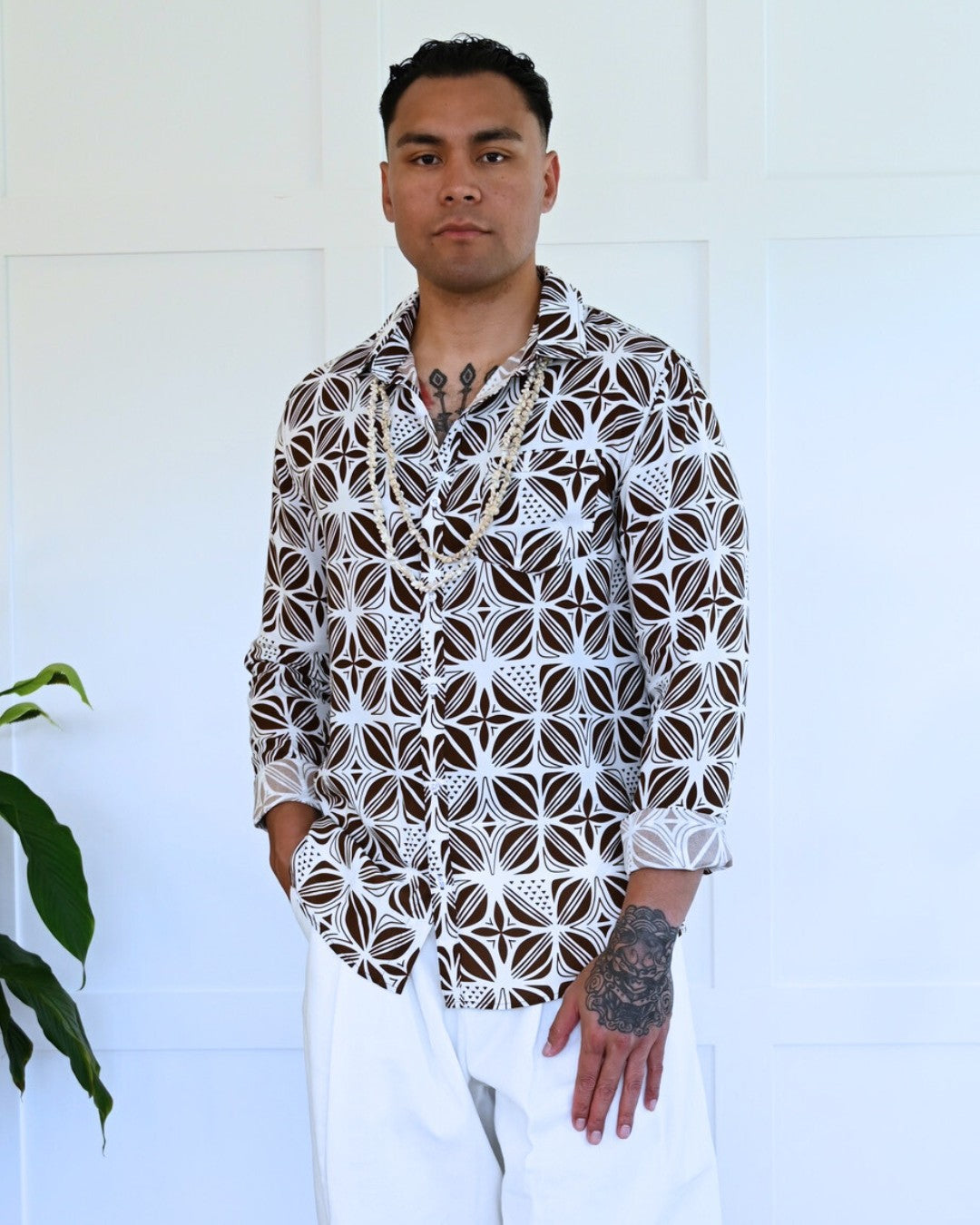 Man wearing a patterned island shirt standing against a white wall with a plant to the side.