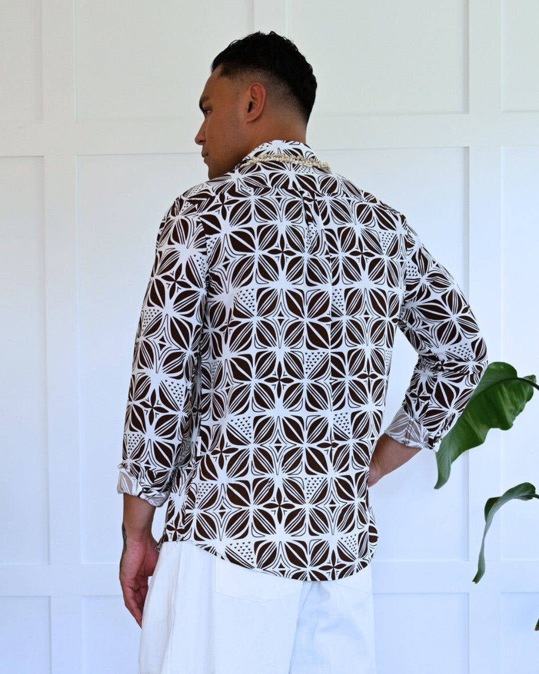 Man wearing a patterned island shirt standing against a white wall with a plant.